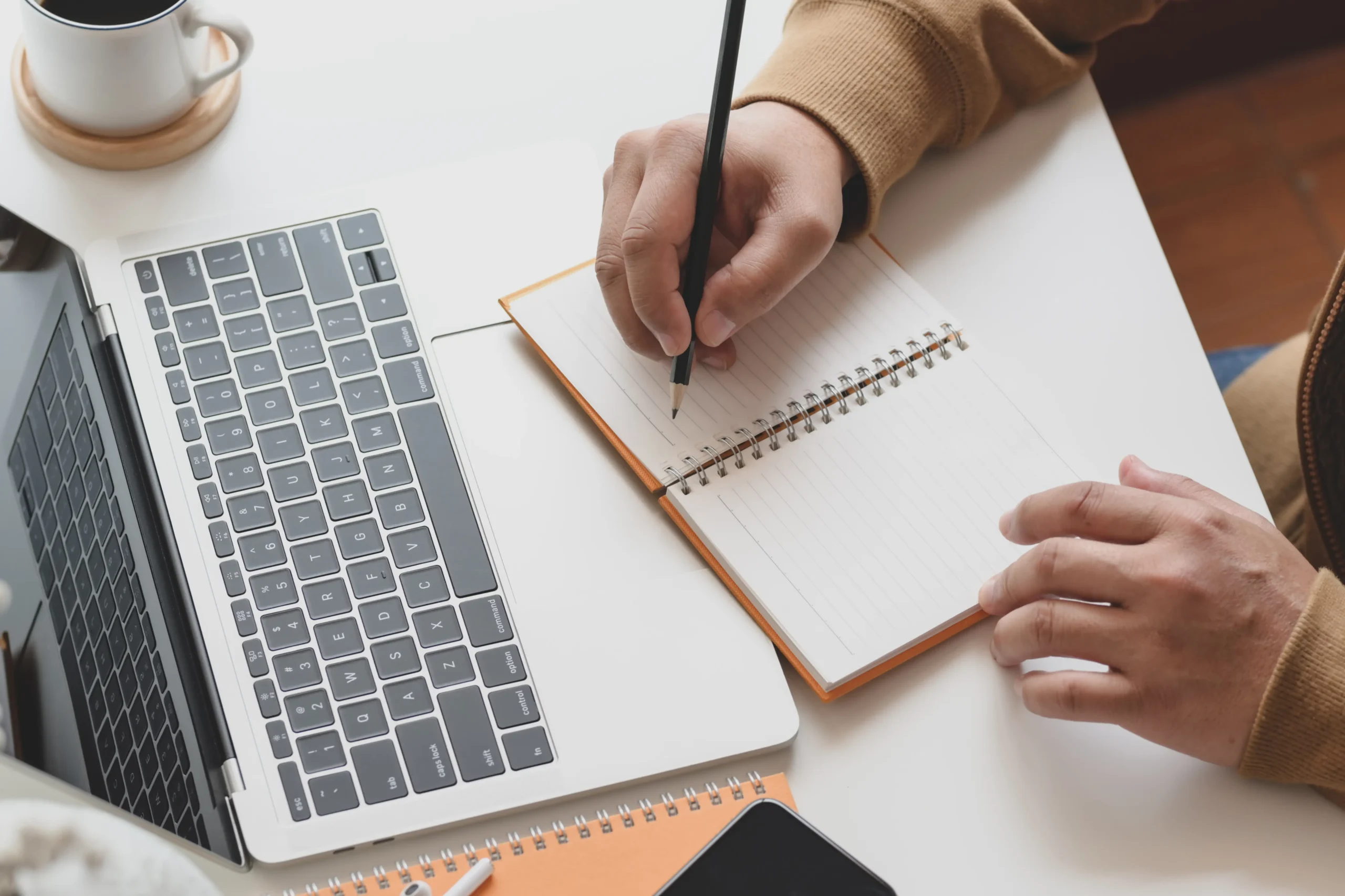 Une personne prend des notes dans un carnet posé devant un ordinateur portable.