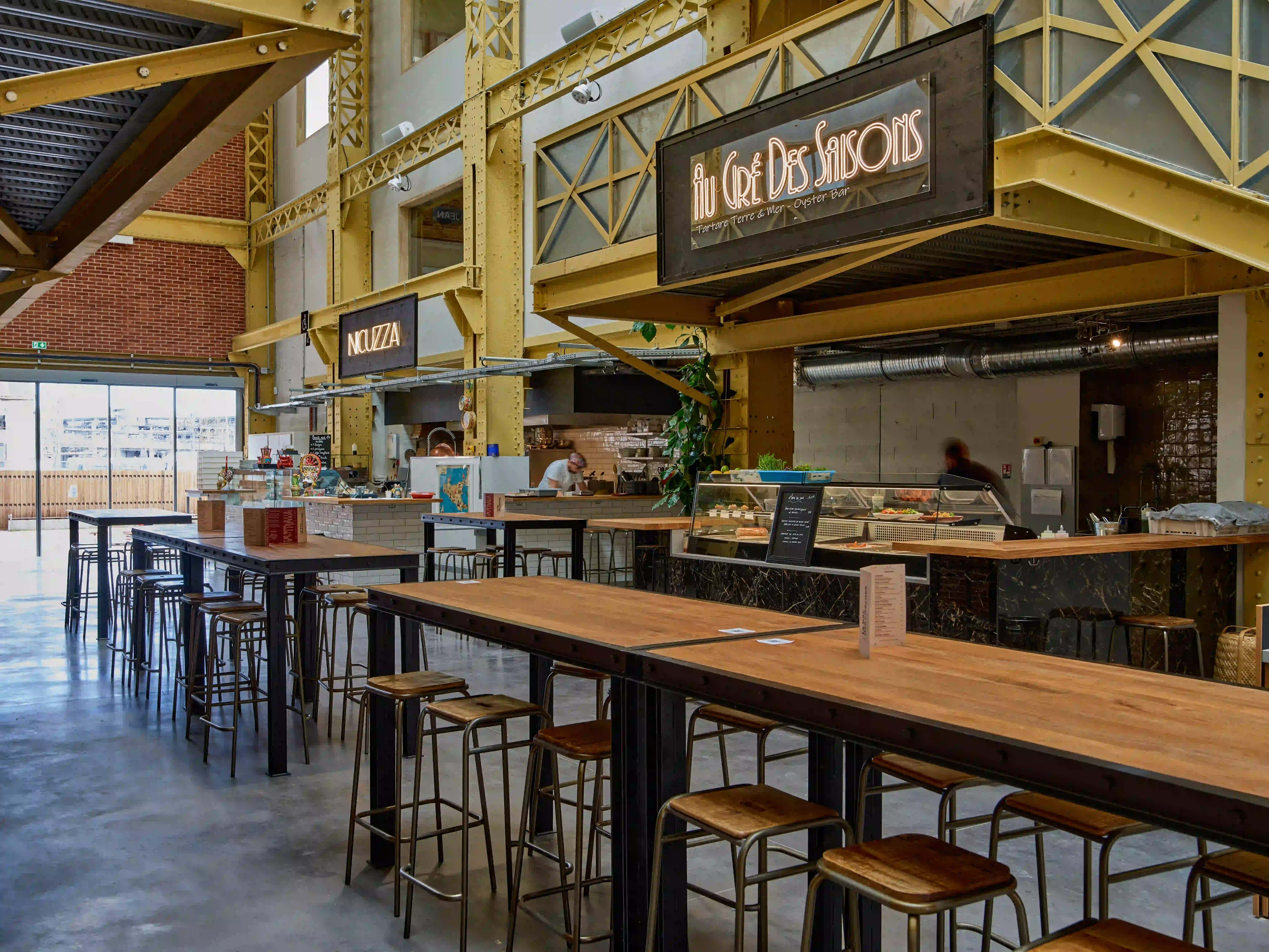 Espace de restauration de l’Atelier des Citernes avec de grandes tables, des comptoirs et une structure métallique jaune.