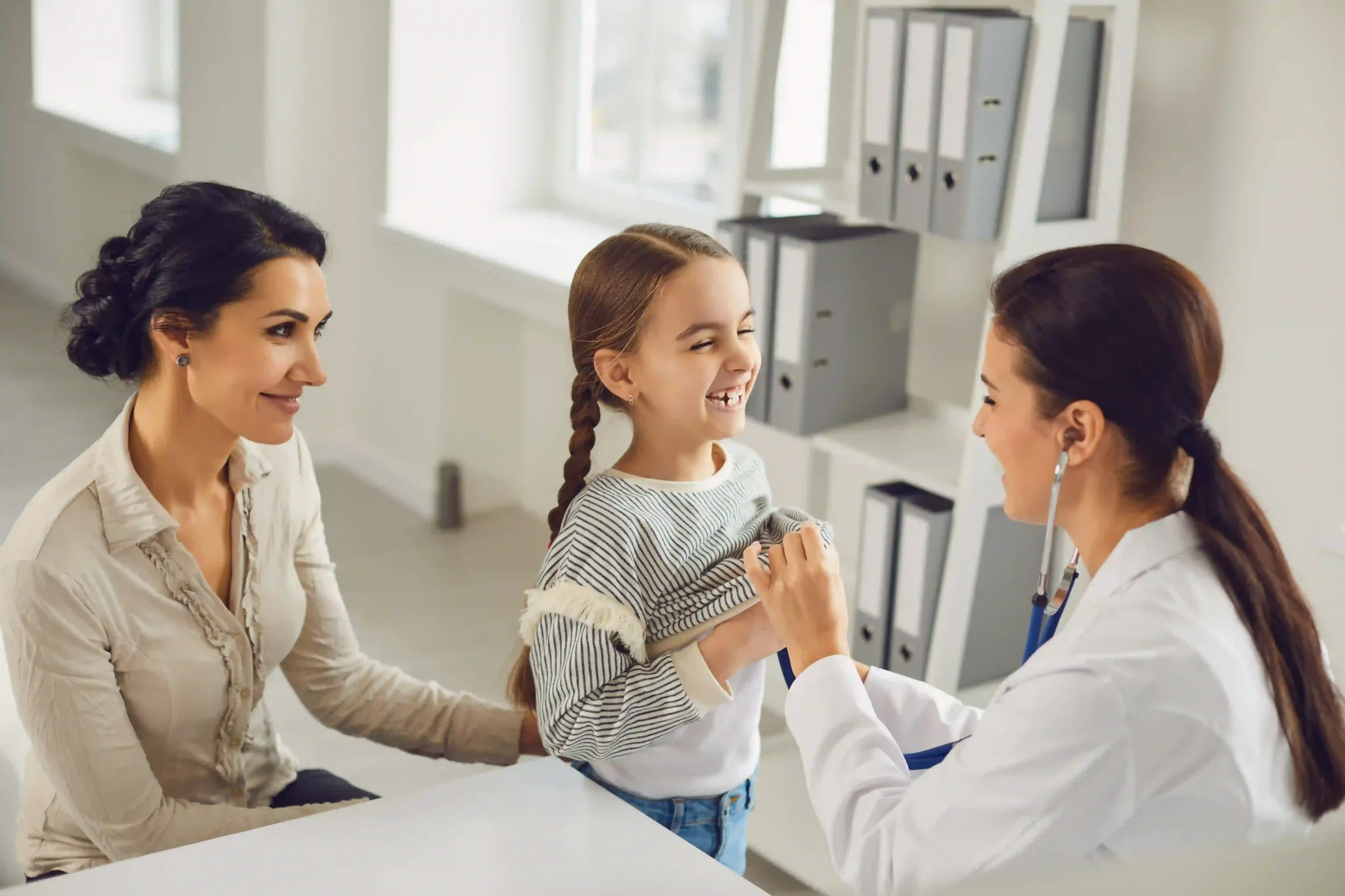 Une pédiatre ausculte une enfant souriante avec un stéthoscope, en présence de sa mère.