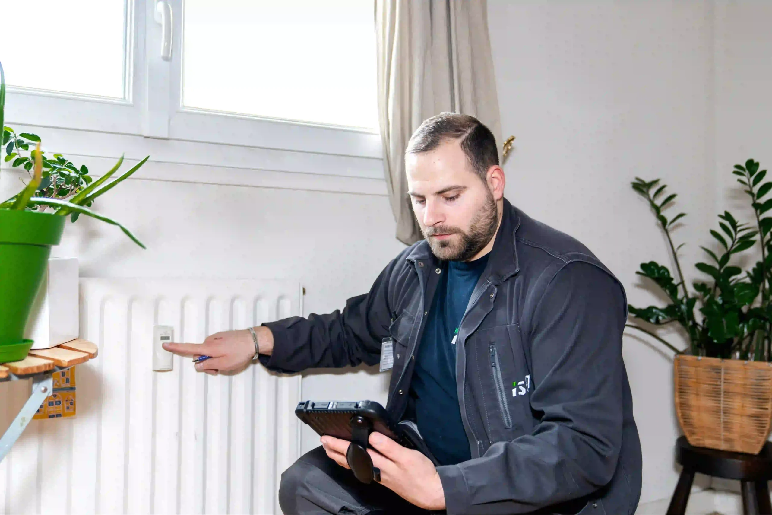 Un technicien ista contrôle un radiateur et son répartiteur de frais de chauffage à l’aide d’une tablette.