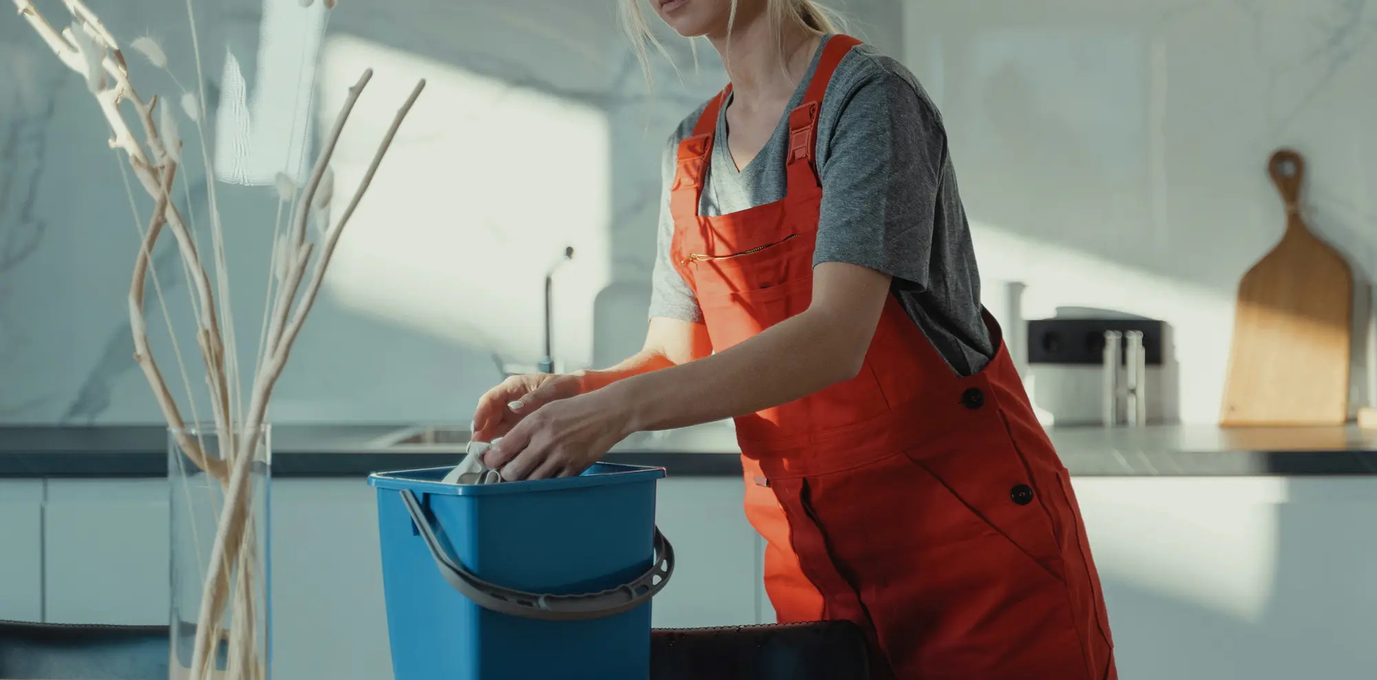 Une professionnelle du ménage essore un chiffon au-dessus d’un seau bleu dans une cuisine.